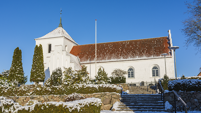 Hvid kirke med rødt tag og tårnspir, omgivet af sneklædte træer og snebeklædt jord.