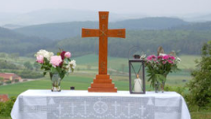 Das Foto zeigt einen aufgebauten Altar auf einer Wiese. Der Tisch trägt ein Kreuz, 2 Blumensträuße links und rechts und eine Kerze.