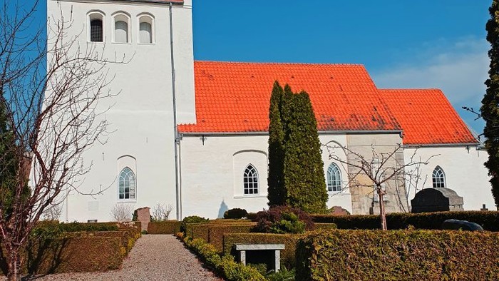 Hyggelig hvid kirke med rødt tag omgivet af veltrimmede hække og en brostensbelagt sti.