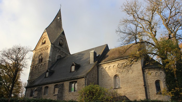 Altes steinernes Gebäude mit spitzem Turm und Rundbogenfenstern, umgeben von Bäumen und Büschen