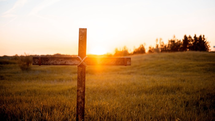 Ein rustikales Holzkreuzschild steht auf einer grasbewachsenen Wiese während eines goldenen Sonnenuntergangs.