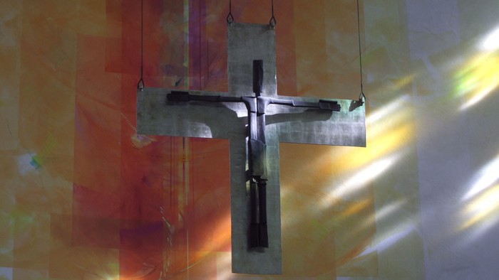 Ein metallenes Kruzifix hängt im Sonnenlicht und wirft lebendige Lichtmuster gegen buntes Kirchenfensterglas.