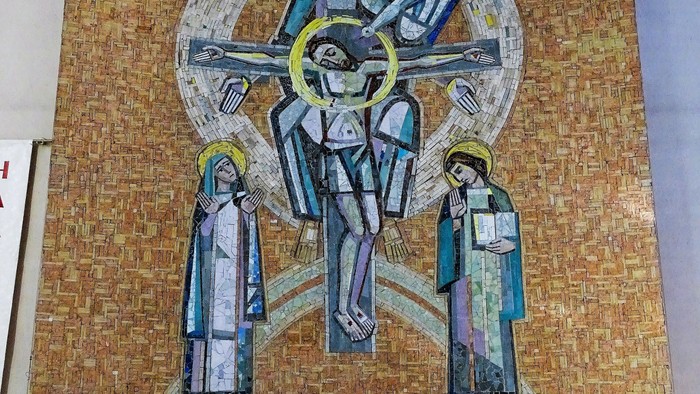 Ein Steinaltar mit Blumenschmuck und zwei Kerzen steht vor einem großen Mosaik-Wandbild. Das Mosaik zeigt zentral die Heilige Dreifaltigkeit.