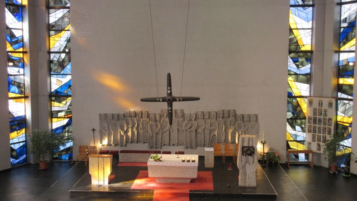 Moderner Kirchenraum der Allerheiligenkirche mit zentralem Steinaltar, einem schwebenden Bronzekreuz und einem großen Steinrelief von Heiligenfiguren im Hintergrund.