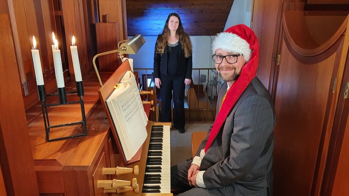 Man in Santa hat plays piano in church with woman standing nearby.