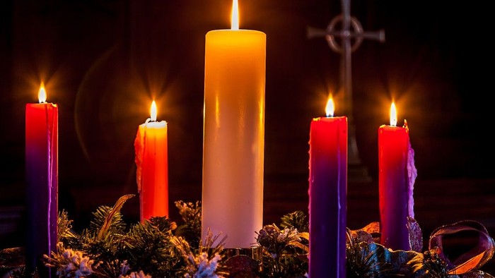 Five lit candles in various colors arranged in a wreath with evergreens and a cross in the background.