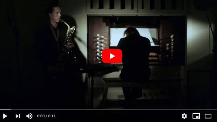 Zwei Personen spielen in einem dunklen Raum Musik, eine am Saxophon, die andere am Klavier.