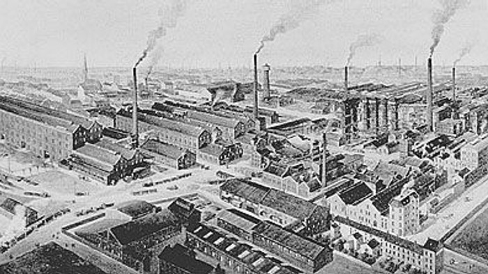 Industrielle Stadtsilhouette mit zahlreichen Gebäuden und Fabrikschloten, aus denen Rauch aufsteigt.