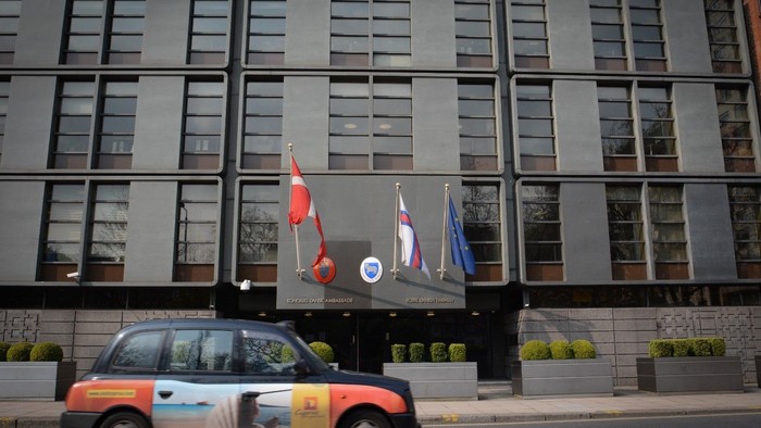 En rød taxi kører forbi et stort, graet kontorbygning med tre flagg.