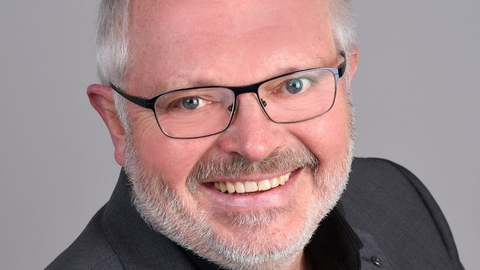 Ein Mann mit Brille und Bart, der lächelt und einen Anzug trägt