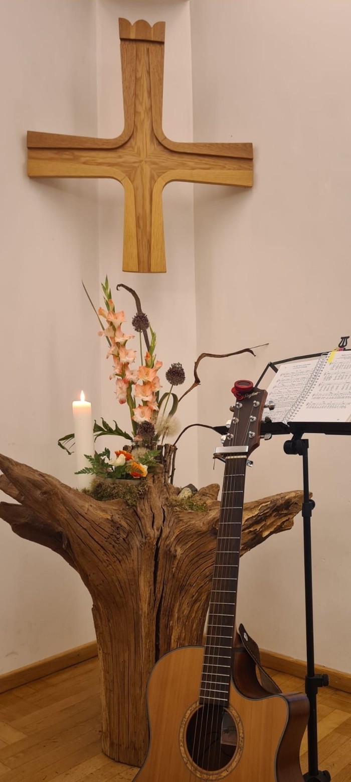 Baumstumpf mit Blumen und brennender Kerze vor großem Holzkreuz, davor Gitarrenhals