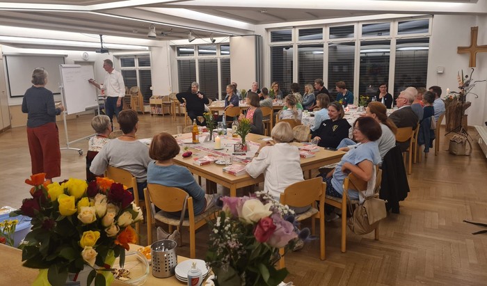 Veranstaltungsraum mit zwei Vortragenden und zuhörenden Menschen an Tischgruppen