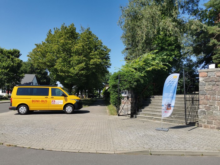 Ein gelber Shuttlebus mit der Aufschrift „BONI-BUS“, geparkt in der Nähe einer Steintreppe mit einem ausgestellten Banner.