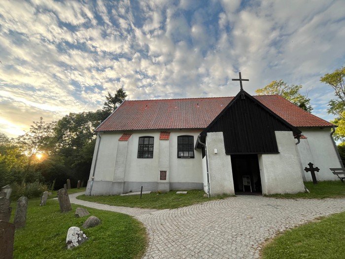 Idyllische alte Kirche mit Kopfsteinpflasterweg und Grabsteinen auf einem ruhigen, sonnenbeschienenen Friedhof.