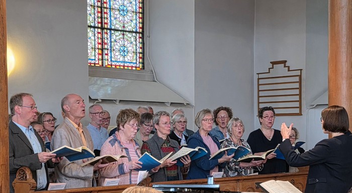 Eine Frau hebt die Hände, um einen Chor unter einem farbigen Kirchenfenster zu dirigieren.