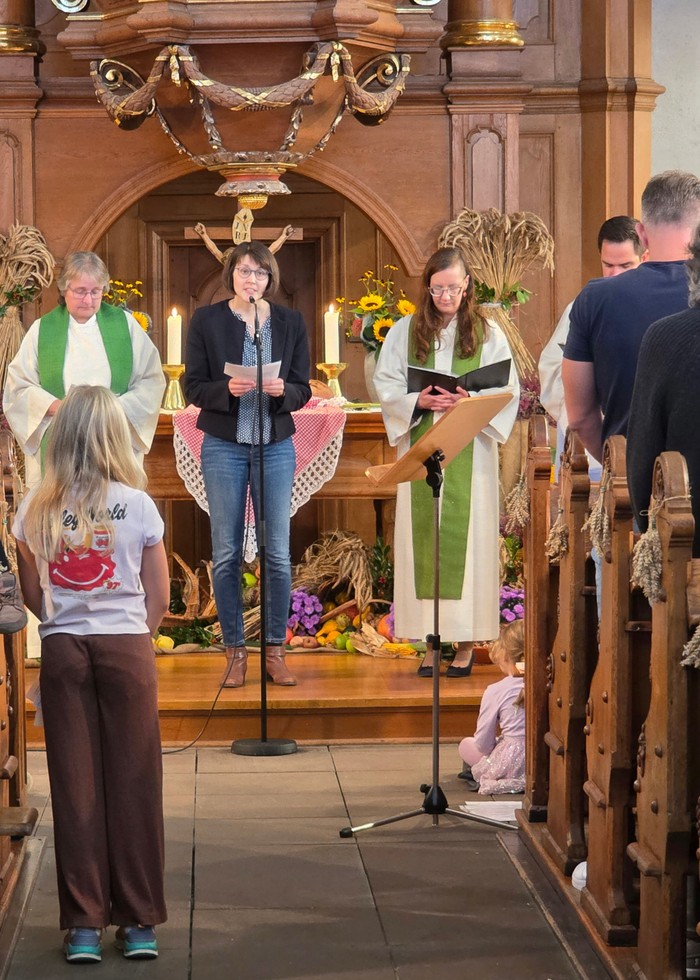 Drei Frauen sprechen in ein Mikrofon vor einem Altar. Aufmerksam hören auch zwei Kinder zu.