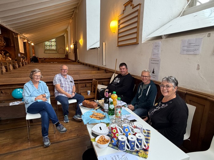 In einer Kirche sitzen mehrere Menschen an einem gedeckten Tisch und lächeln in die Kamera.