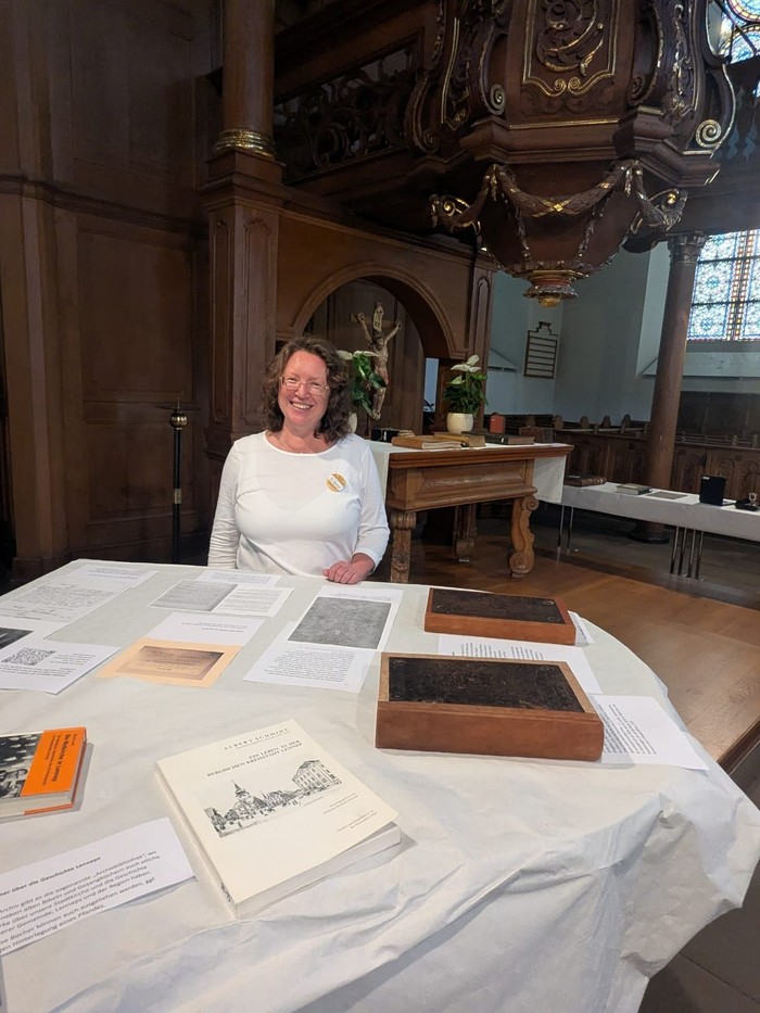 Eine Frau mit weißem T-Shirt sitzt an einem Tisch mit Büchern und Computerausdrucken vor einem Altar.