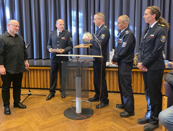 Ein Mann bekommt von vier Feuerwehrleuten ein Geschenk. In der Mitte ein Rednerpult. 