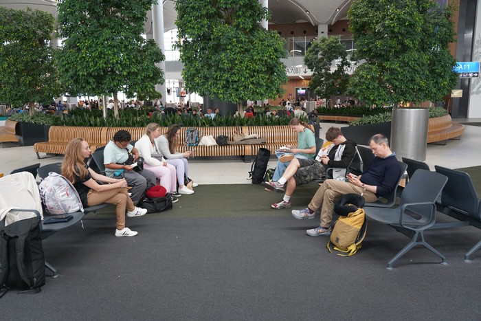Mehrere Menschen sitzen am Flughafen im Wartebereich. Im Hintergrund sind Bäume zu sehen
