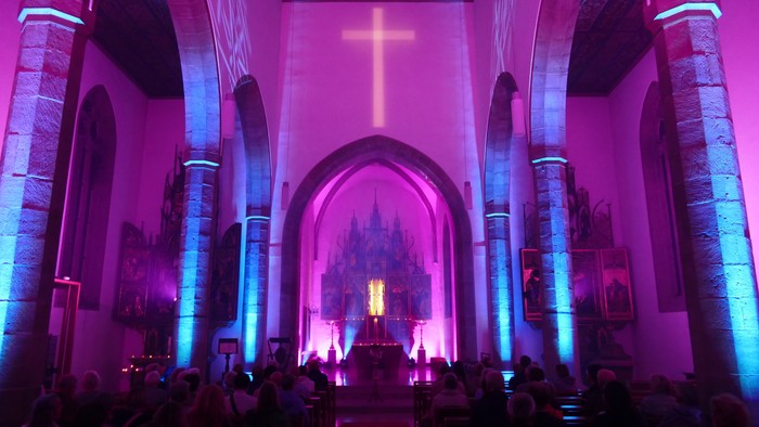 Lebhaft beleuchteter Kircheninnenraum mit versammelter Gemeinde unter einem großen, erleuchteten Kreuz während eines Gottesdienstes.