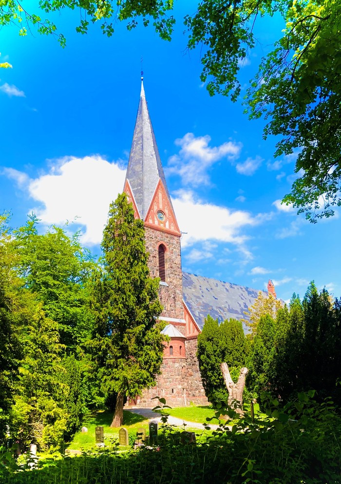 Ein hohes, spitzes Kirchenturm aus rotem Backstein, umgeben von üppigem Grün und einem klaren blauen Himmel