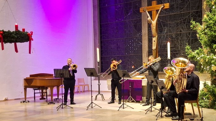 Ein Bläserchor spielt in einem Altarraum. Rechts hängt ein Adventskranz. Am linken Bildrand ein Weihnachtsbaum.