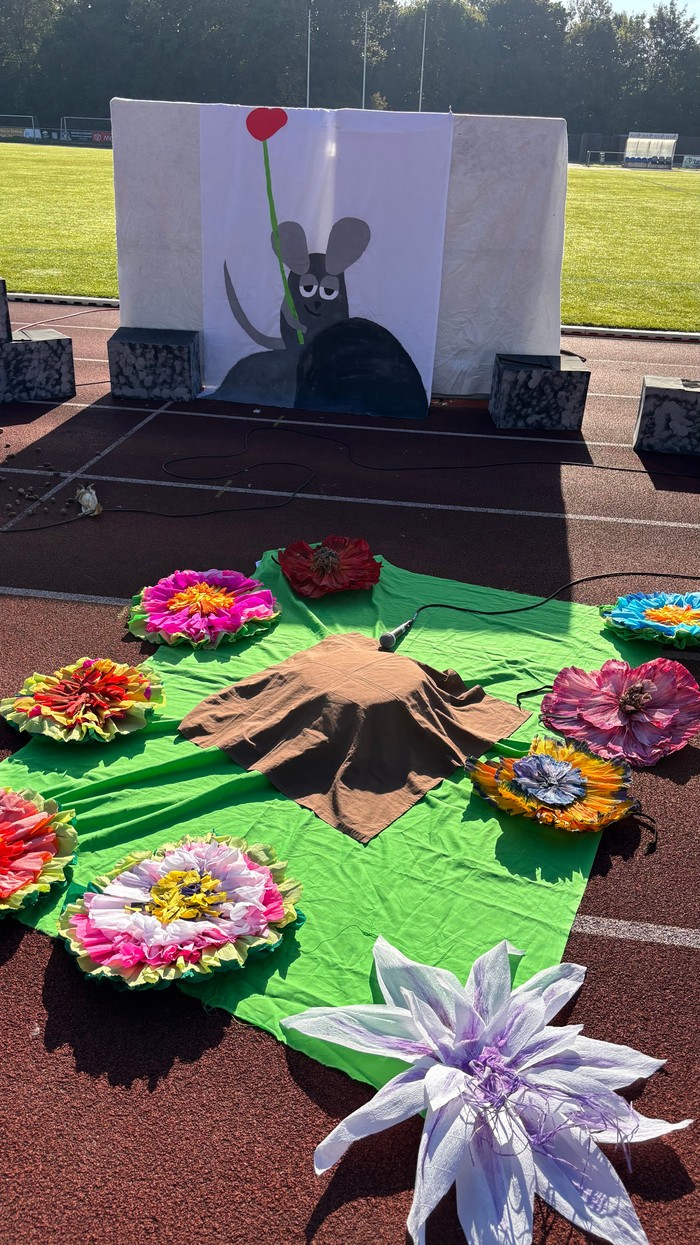 Man sieht ein Bühnenbild. Im Hintergrund ein Bild aus der Geschichte von Frederick. Vorne gebastelte Blumen und in der Mitte einen Baumstamm. Das ganze ist auf der Laufbahn des Stadions aufgebaut. Hinter der Bühne sieht man den Rasen des Stadions.