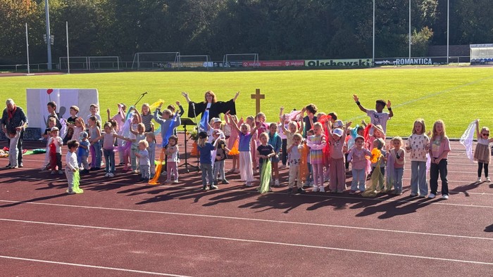 Auf der Tartanbahn des Jahnstadions stehen viele singende Kinder vor einem aufgebauten Altar. Sie singen zusammen mit Pfarrerin Annika Wilinski ein Lied im Erntedankgottesdienst 2027.