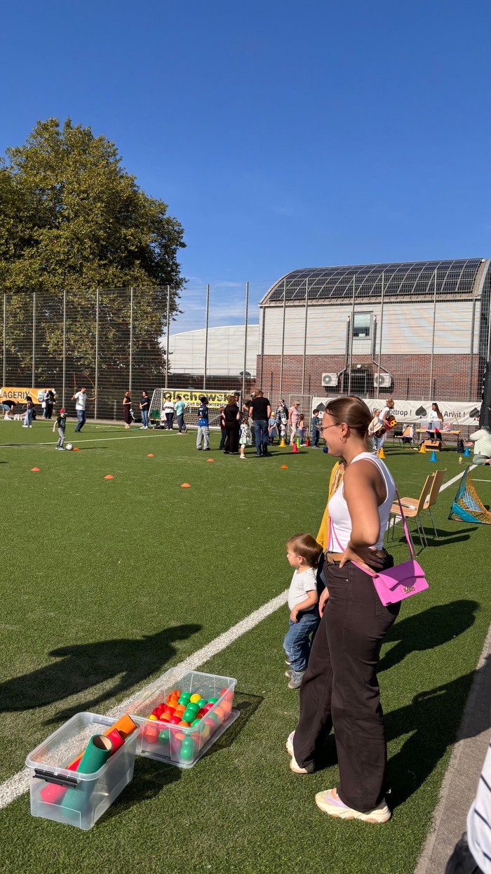 An sieht einen Ausschnitt vom Kunstrasenplatz neben dem Jahnstadion. Dort sind verschiedene Sportstationen aufgebaut. Ein Slalomparkour sowie Fußball-Darts ist zu sehen. An den Stationen sieht man beschäftigte Kinder und zuschauende Erwachsene.