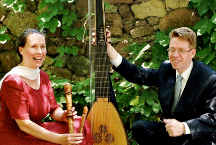 Zwei Personen spielen im Freien traditionelle Instrumente in der Nähe einer Steinmauer und lächeln sich an.