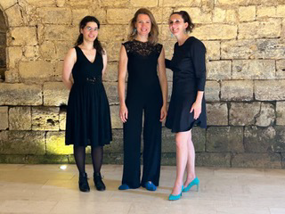 Drei Frauen in eleganter schwarzer Kleidung und türkisfarbenen High Heels posieren gemeinsam vor einer Steinmauer.
