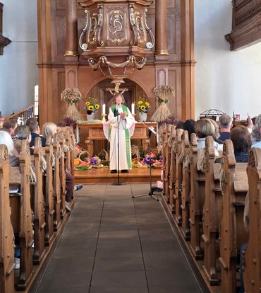 Eine Frau mit weißem Talar und grünem Tuch steht vor einem Altar über ihr die hölzerne Kanzel und spricht zu den Menschen in der Kirche.