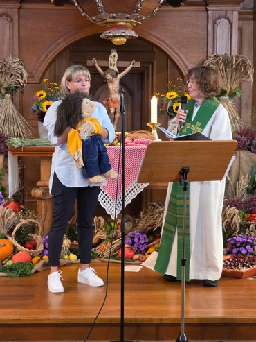 Eine Pfarrerin im weißen Talar und grünem Bandd hält ein Mikrofon vor einem mit Erntedank-Früchten geschmückten Altar zu einer Sprechpuppe in der Hand einer Frau.