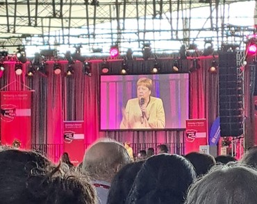 Auf einem großen Bildschirm spricht eine Frau mit gelben Jacket, die ein Mikrofpn in der hand hält. Auf einem großen Bildschirm spricht eine Frau mit gelben Jacket, die ein Mikrofpn in der hand hält.