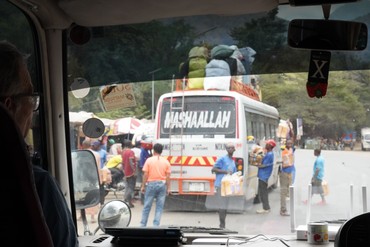 Blick aus einem Fahrzeug auf einen Kleinbus mit zahlreichen Gepäckstücken auf dem Dach. Rundherum stehen Menschen und Verkaufsstände an einer belebten Straße.