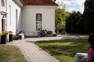 Eine kleine Gruppe sitzt bei sonnigem Wetter auf einer Bank vor der Dorfkirche Alt-Staaken und schaut gemeinsam in ein Bilderbuch – eine der Stationen der Kirche Kunterbunt.