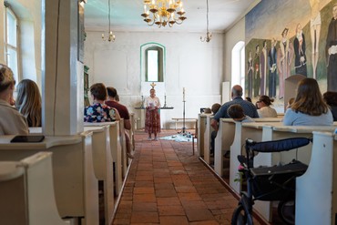 Blick durch den Mittelgang der Dorfkirche Alt-Staaken während der Kirche Kunterbunt. Eine Mitarbeiterin steht vor dem Altar und spricht zu den zahlreichen Kindern und Erwachsenen in den Kirchenbänken.