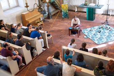 Kinder und Erwachsene sitzen während der Kirche Kunterbunt in den Kirchenbänken. Vor dem Altar erzählt eine Mitarbeiterin eine Geschichte, vor ihr liegen Figuren und Materialien auf einer Decke.