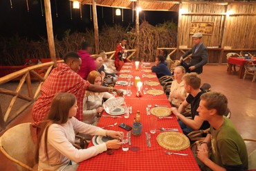 Die Reisegruppe sitzt abends an einer langen, rot gedeckten Tafel in einem offenen Holzpavillon. Eine Person in traditionell rot kariertem Gewand schenkt Wasser ein.
