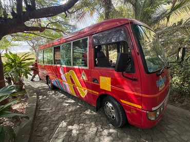 Roter Kleinbus mit bunten Grafiken, der auf einem gepflasterten Weg unter großen Bäumen und tropischer Vegetation steht. 