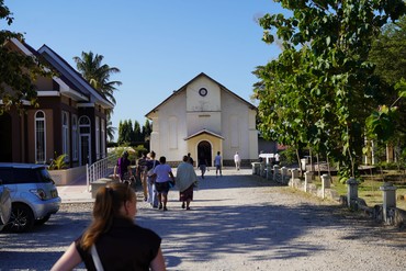 Blick auf die Kirche in Tanga, zu der eine Gruppe von Besucherinnen und Besuchern über das Kirchengelände läuft. 