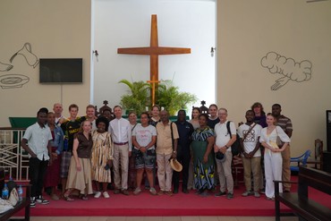 Gemeinsames Gruppenfoto von tansanischen und deutschen Jugendlichen und Erwachsenen vor dem Altarraum der Kirche in Tanga mit einem großen Holzkreuz im Hintergrund.