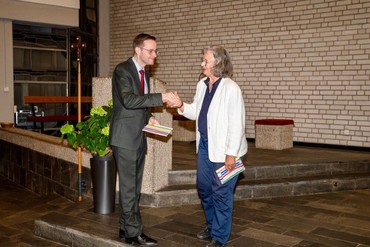 Zwei Personen auf einer Plattform bei einer feierlichen Übergabe, mit Kreuz und Pflanze im Hintergrund.