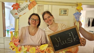Zwei Personen stehen hinter einem Bilderrahmen. Auf eine Tafel haben sie geschrieben wofür sie dankbar sind: Musik