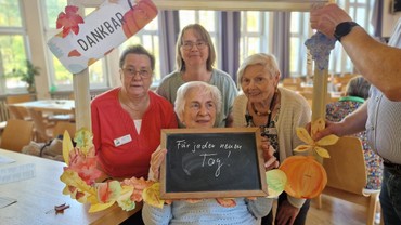 Hinter einem Bilderrahmen stehen vier Personen. Auf eine Tafel haben sie geschrieben wofür sie dankbar sind: jeden neuen Tag!