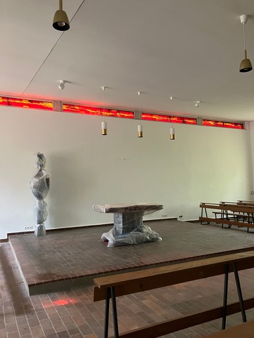 Moderner Galerieraum mit abstrakten Skulpturen und einer rot beleuchteten Wandpräsentation, minimalistischen Tischen und Bänken.