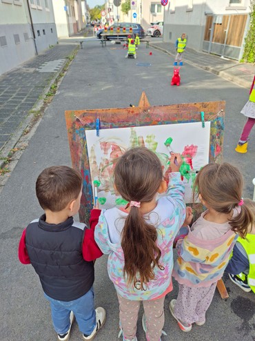 Ein Junge und zwei Mädchen, alle drei im Kita-Alter, malen an einer Kinderstaffelei mit Pinseln an einem Straßen-Bild.